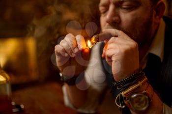 Portrait of bearded man lights a cigar with a match, closeup view. Tobacco smoking culture, specific flavor. Male smoker leisures in office