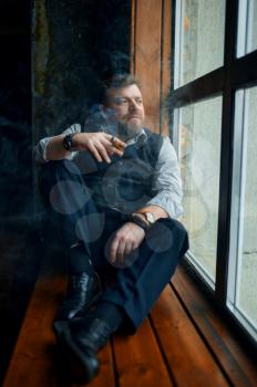 Man sitting on the windowsill and smokes a cigar, vintage office interior on background. Tobacco smoking culture, specific flavor. Male smoker looks at the window