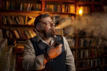 Serious bearded man smokes a cigar in office, bookshelf on background. Tobacco smoking culture, specific flavor. Male smoker leisures