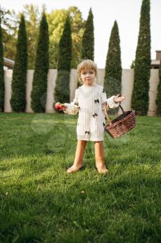 Little girl with basket eats an apple in the garden. Female child poses on the lawn on backyard. Kid having fun on playground outdoors, happy childhood