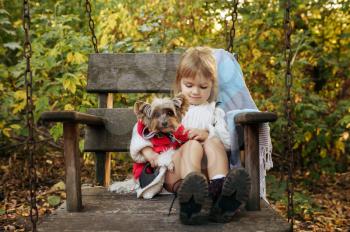 Kid with funny dog are sitting in big wooden chair in the garden. Female child with puppy poses on backyard. Little girl and her pet having fun on playground outdoors, happy childhood