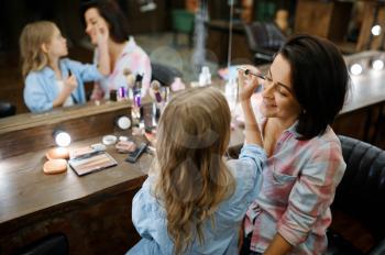 Adorable daughter applies mascara for eyelashes to her mother in makeup salon. Mom and little girl play stylists together, happy childhood, glamour family