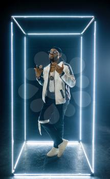 Stylish rapper in sunglasses poses in illuminated cube, dark background. Hip-hop performer, rap singer, break-dance performing, entertainment lifestyle