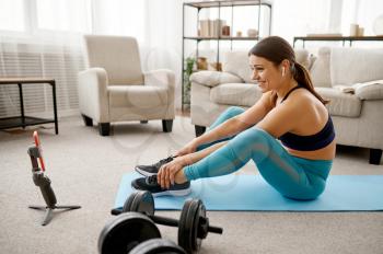 Smiling girl sits on the floor at home, online fitness training at the laptop. Female person in sportswear, internet sport workout, room interior