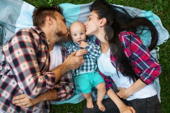 Mother and father kissing their little baby, top view. Mom and dad with male kid on lawn, parents on picnic with child in the forest, family happiness