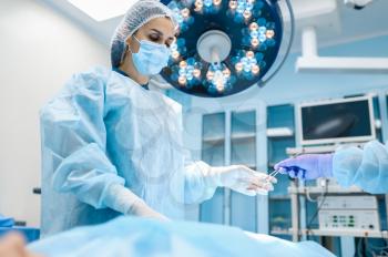 Female surgeon in gown and mask in operating room, lamp on background, surgery operation. Doctor in uniform, medical clinic worker, medicine and health