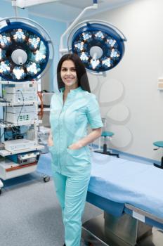 Smiling female surgeon poses at the operating table, lamp on background, surgery. Doctor in uniform, medical clinic worker, medicine and health, professional healthcare in hospital