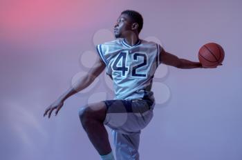 Basketball player with ball shows his skill in studio, jump in action, neon background. Professional male baller in sportswear playing sport game, tall sportsman