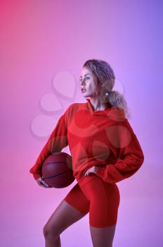 Sexy female athlete in red hoodie poses with ball in studio, neon background. Fitness sportswoman at the photo shoot, sport concept, active lifestyle motivation
