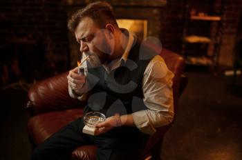 Portrait of bearded man with ashtray lights a cigarette, closeup view. Tobacco smoking culture, specific flavor. Male smoker leisures in office