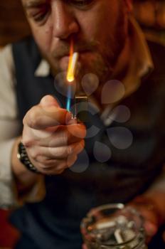 Portrait of bearded man with ashtray lights a cigarette, closeup view. Tobacco smoking culture, specific flavor. Male smoker leisures in office
