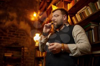 Bearded man smokes cigarette with mouthpiece, bookshelf and rich office interior on background. Tobacco smoking culture, specific flavor