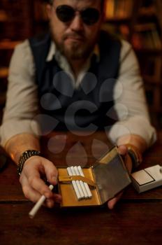 Serious man in sunglasses holds cigarette case, bookshelf and rich office interior on background. Tobacco smoking culture, specific flavor. Smoke habit