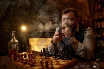 Man smokes a cigar, drinks alcohol beverage and play chess, bookshelf and vintage office interior on background. Tobacco smoking culture, specific flavor. Male smoker leisures at the chessboard
