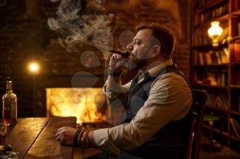 Man smokes a cigar and drinks alcohol beverage, bookshelf and rich office interior on background. Tobacco smoking culture, specific flavor