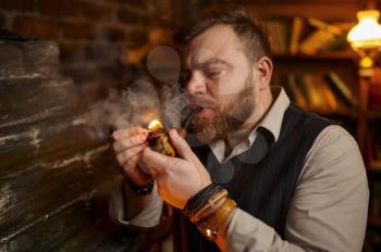 Portrait of bearded man lights smoking pipe with a match, closeup view. Tobacco smoking culture, specific rich flavor. Male smoker leisures in office