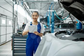 Female mechanic with laptop checks engine, car service, professional diagtostic. Vehicle repairing garage, woman in uniform, automobile station interior on background