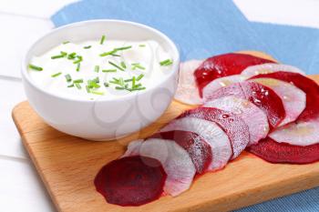 bowl of white yogurt with thin slices of beetroot and white radish on wooden cutting board