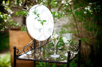 Little table, mirror and glasses in bushes of a cherry. Spring garden.
