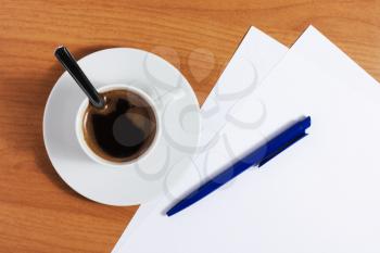 A cup of coffee on a wooden table with blank papers and blue pen, top view