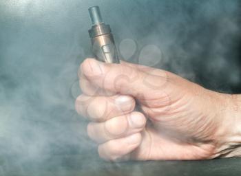 Hand with an electronic cigarette in the clouds of steam on a dark background