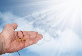 Hand with a wooden cross against the blue sky with clouds and sun rays