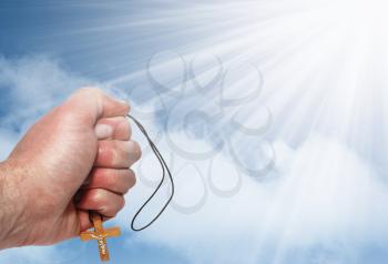 Hand with a wooden cross against the blue sky with clouds and sun rays