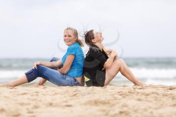 Two beautiful smiling woman sitting with his back to each other and resting on the sand on the seashore. Shallow depth of field. Focus on the models.