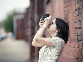 Pretty young woman taking photo outdoors. Girl holding vintage camera and taking photographs. Retro style photo. Selective focus on woman. Toned photo with copy space.