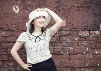 Smiling young woman in a white blouse and a hat posing against vintage brick wall background. Vintage portrait of a woman. Toned photo with copy space. Vintage style photo.