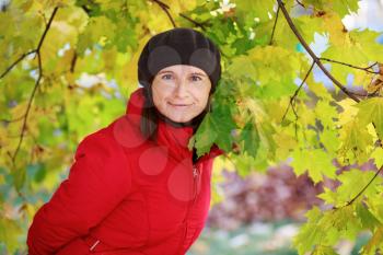 Girl in a beret and red jacket on the background of green and yellow autumn maple leaves. Woman looking into the camera. Selective focus on model.