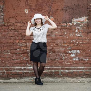 Photo of a woman in a white hat, blouse and black skirt, standing against the backdrop of an old vintage brown brick wall. Model holds the edges of the hat by hand. Portrait of a woman in a retro styl