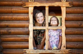 Happy smiling mom and daughter look out of the window a wooden timbered hut. Mother and baby girl looking out the window. Happy family.