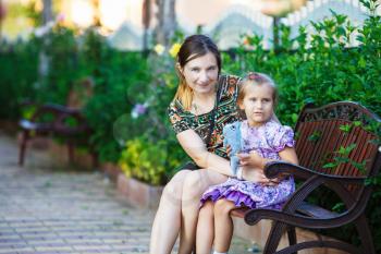 Mother and little daughter sit on a park bench on a background of bright green foliage. Hugging mother and daughter. Family portrait. Selective focus.