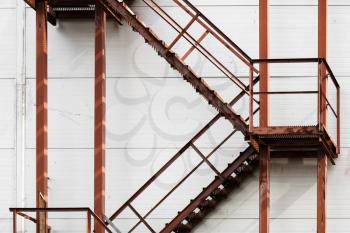 Old rusty metal ladder against a white wall background. Metal steps with handrails and staircases covered with rust. Facade of industrial buildings.