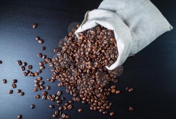Coffee beans scattered from a canvas bag against black wooden table background.
