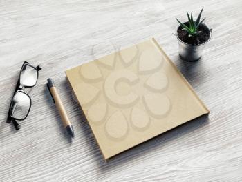 Photo of closed blank sketchbook, glasses, pen and plant on light wood table background. Blank stationery template.
