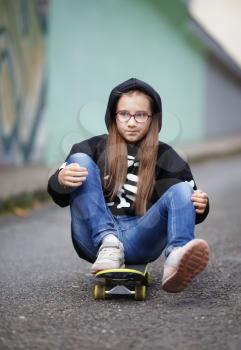 Girl rides on skateboard. Child in a black sweatshirt sitting on her skateboard. Vertical shot. Selective focus.