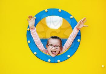 Happy girl in blue porthole on yellow background. Child has fun on the playground.