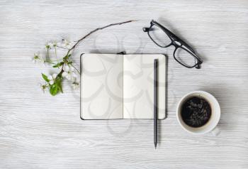 Stationery set. Blank notebook, coffee cup, pencil, glasses and flower on light wood table background. Flat lay.
