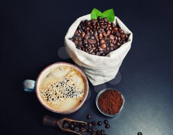 Photo of coffee cup, roasted coffee beans in canvas bag and ground powder on black kitchen table.
