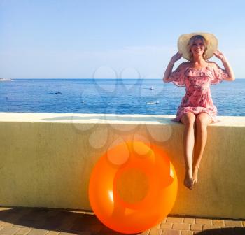 A woman with a beautiful figure is sitting on a concrete fence in front of the sea. Nogshinahschiny orange inflatable circle
