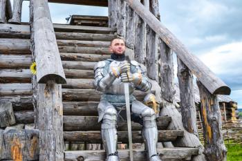 Knight in the armor on the wooden steps. Knightly armor and weapon. Semi - antique photo.