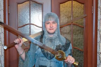 A man dressed up in a knight's hauberk at home in his room. Knight's sword in the hands of a man. Knightly chain armor and sword. Fitting of a knightly armor.