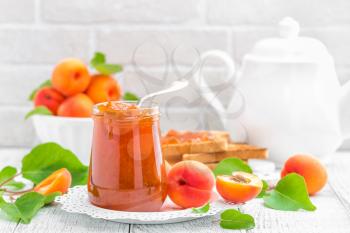 Apricot jam in a jar and fresh fruits with leaves on white wooden table, breakfast