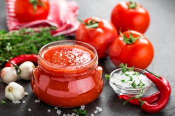 Tomato paste, puree in glass jar and fresh tomatos on dark background. Hot vegetable sauce with chili pepper ant tomatoes