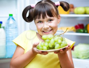 Happy girl eating grapes standing near refrigerator with fruits and vegetables