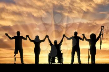Invalids on crutches and in wheelchair and prosthesis in foot and healthy people standing on sunset background and holding hands. Concept happy disabled