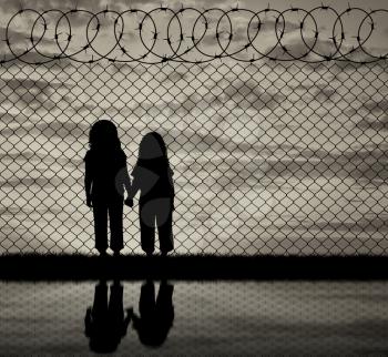 Concept of the refugees. Silhouette of hungry children in desperate refugees near the fence at sunset and reflection