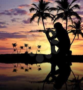 Silhouette of a terrorist with a weapon against a background of a sunset with palm trees and reflection in water